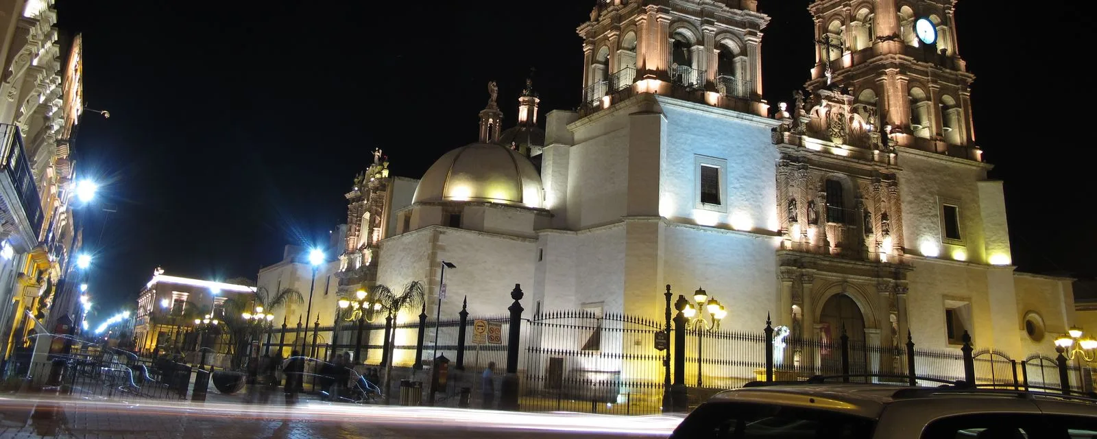 La Catedral Basílica de Durango iluminada de noche, con su cúpula y su torre de cantera sobre la calle y las farolas encendidas de la plaza