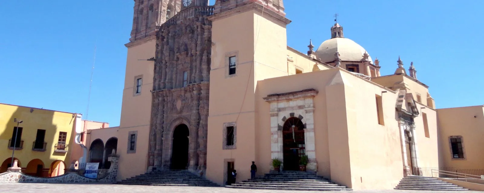 La parroquia de Nuestra Señora de los Dolores vista de tres cuartos desde la plaza, con su torre labrada y su cúpula blanca contra el cielo despejado