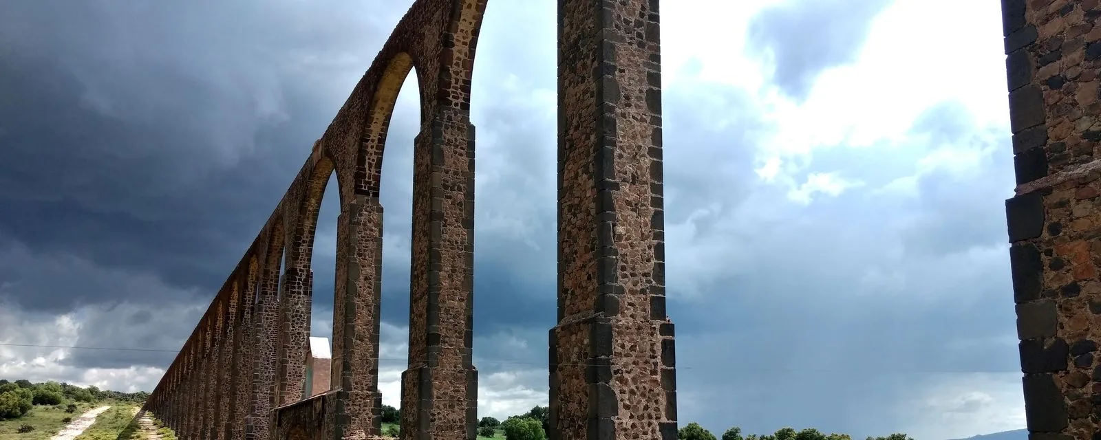 La arquería del Acueducto del Padre Tembleque vista desde abajo: pilares de mampostería y arcos de medio punto alejándose en perspectiva bajo un cielo de tormenta
