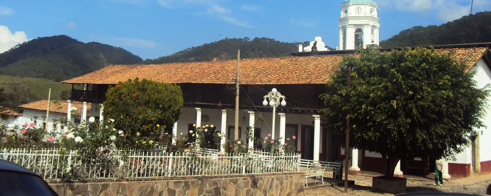 La plaza de San Sebastián del Oeste: el portal de tejado rojo y columnas blancas, la torre del reloj asomando detrás y las montañas de la sierra al fondo