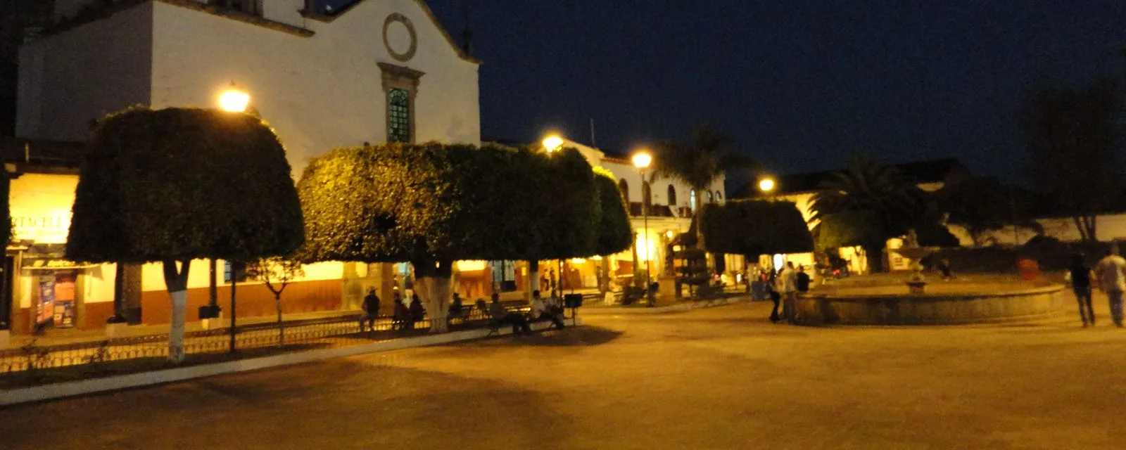 La plaza de Tacámbaro al anochecer: los laureles podados en cubo iluminados desde abajo y la fachada blanca del templo al fondo