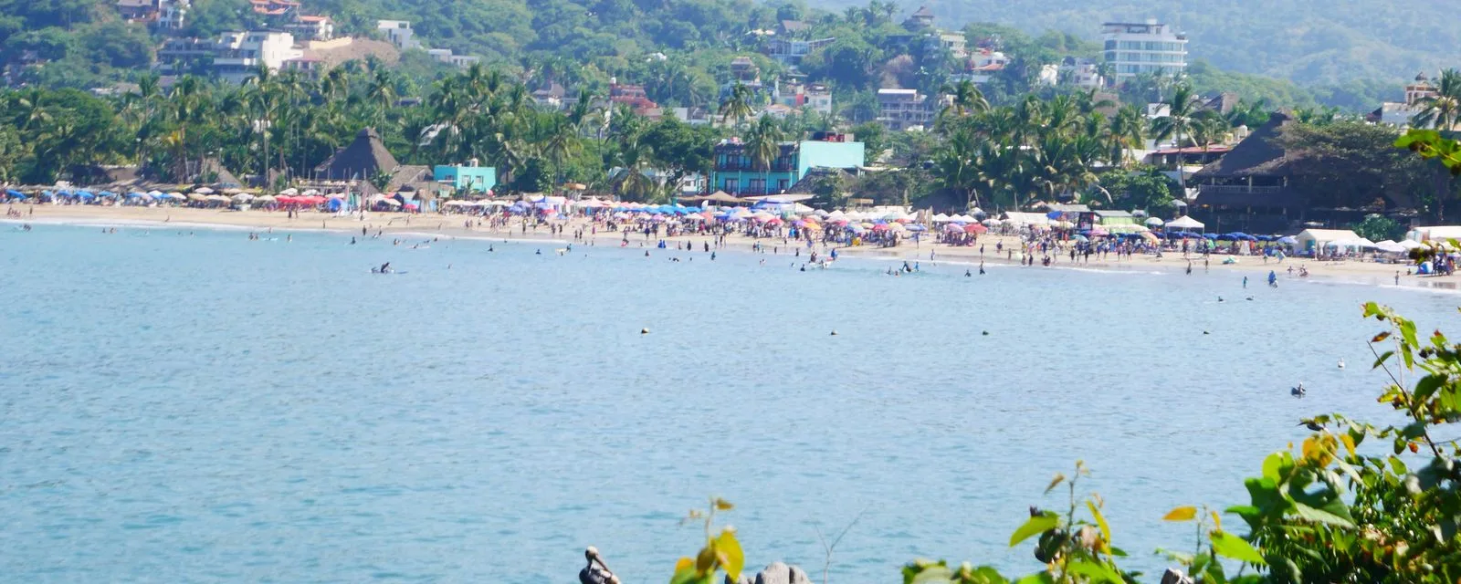 La bahía de Sayulita vista desde el extremo del arco: la playa llena de sombrillas y palapas, con el pueblo trepando la ladera verde detrás