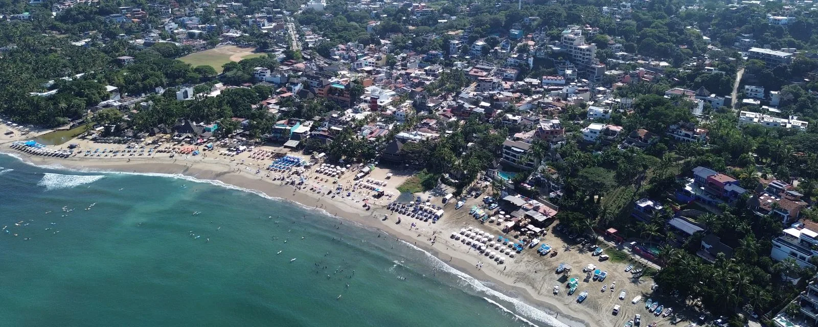 Sayulita desde el aire: la bahía con las sombrillas alineadas en la arena, el oleaje entrando y el pueblo entre la vegetación