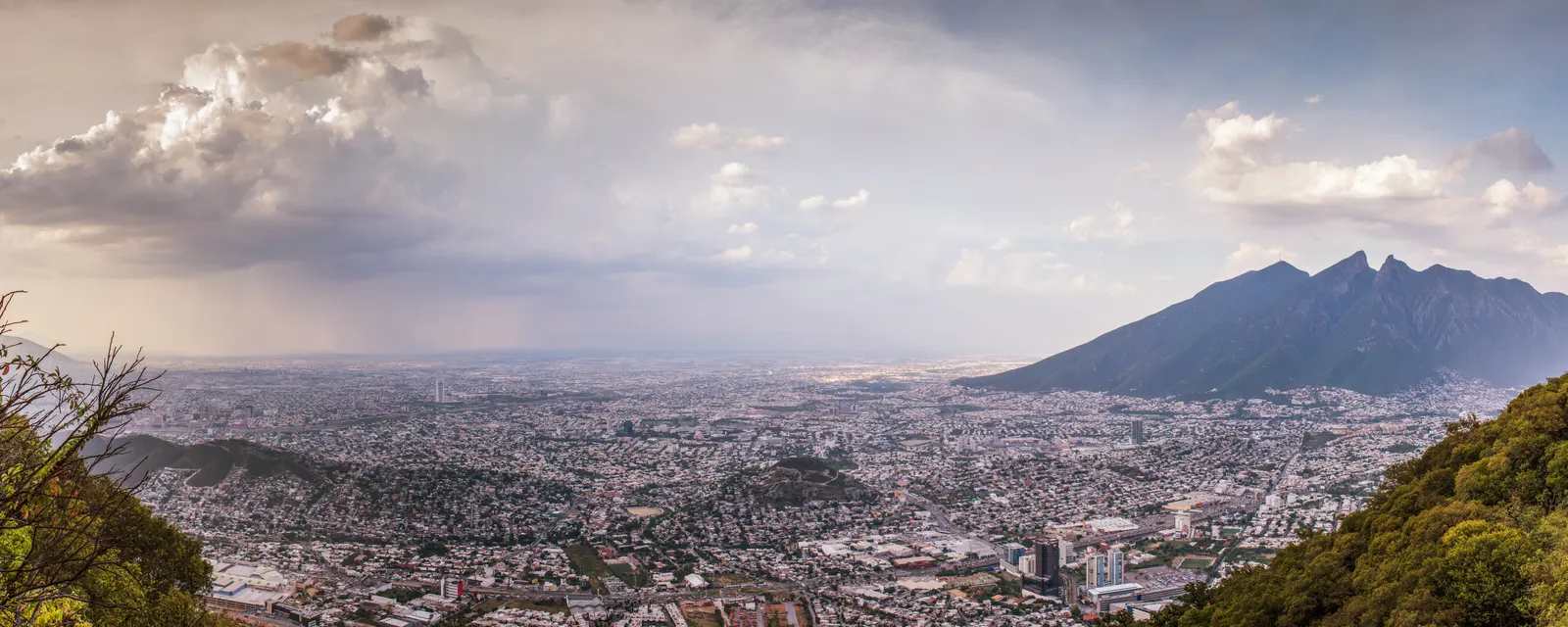 Monterrey vista desde la sierra: la mancha urbana llenando el valle bajo una tormenta, con el perfil de silla de montar del Cerro de la Silla a la derecha
