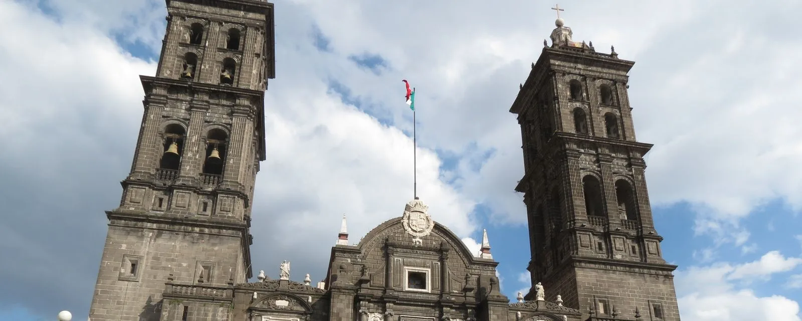 Las dos torres de cantera gris de la Catedral de Puebla recortadas contra un cielo de nubes, con la bandera ondeando entre ellas