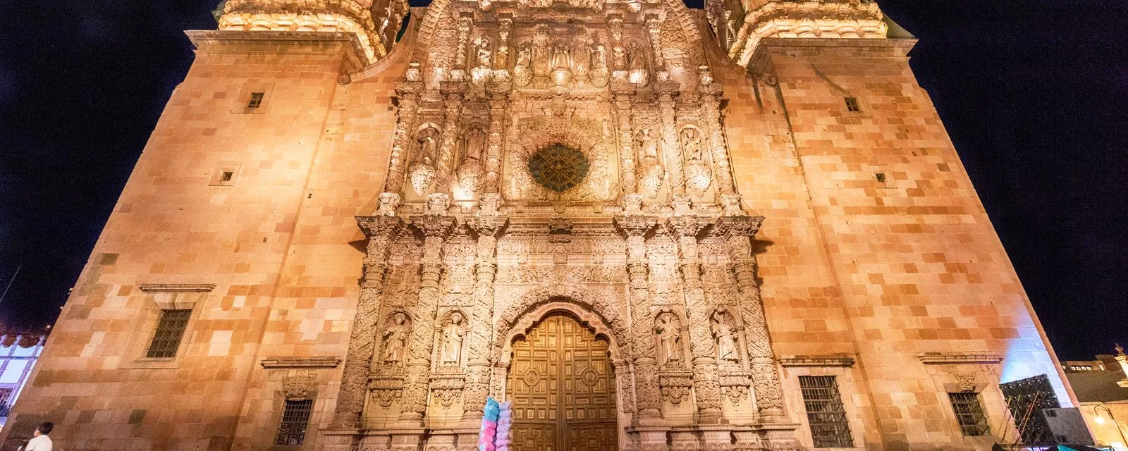 La fachada principal de la Catedral Basílica de Zacatecas iluminada de noche: cantera rosa labrada de arriba abajo con figuras, columnas y un rosetón al centro