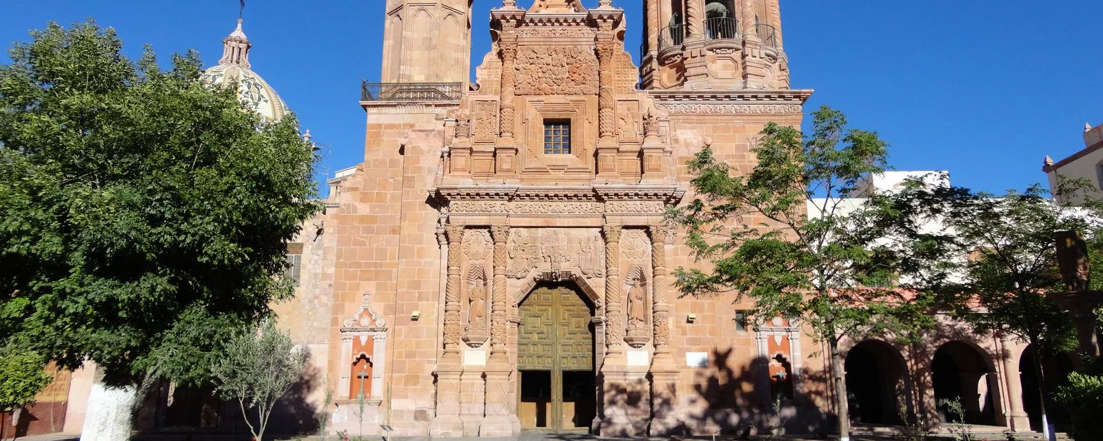La portada de cantera labrada del templo de Guadalupe, Zacatecas, con su torre y la cúpula del convento asomando detrás de los árboles del atrio