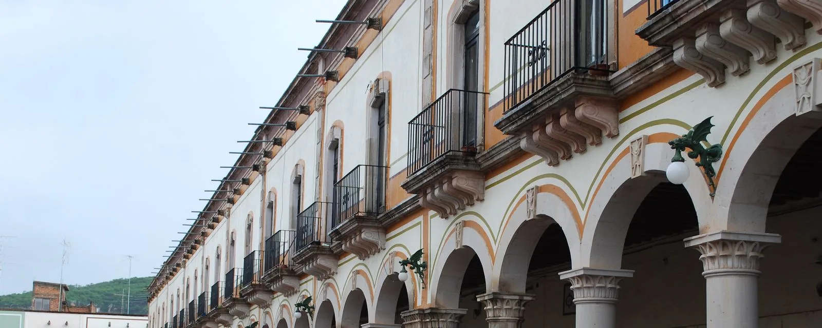Los portales del Teúl de González Ortega: arcada de dos niveles con balcones de herrería y cenefas pintadas, y el cerro asomando al fondo